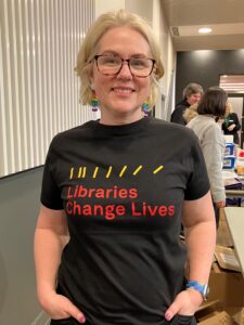 Headshot of a smiling blonde woman wearing black-rimmed glasses and a black t-rhist with yellow lines and the words 'Libraries Change Lives' in red written on it.